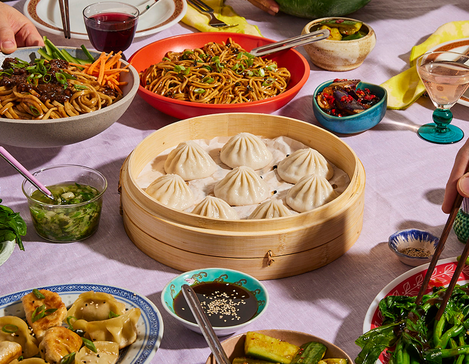 Basket of steamed soup dumplings in a bamboo steamer on a table surrounded by noodles, pan-fried dumplings, sauces, and side dishes