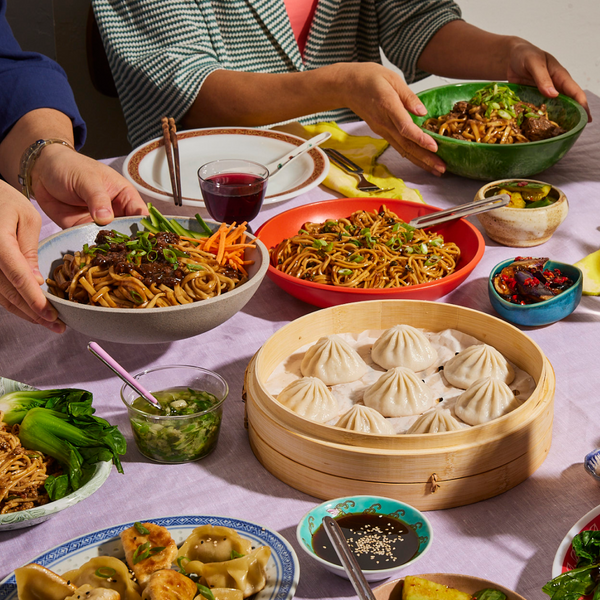 Two people serving bowls of noodles around a table with a bamboo steamer of soup dumplings, dipping sauces, and vegetable sides