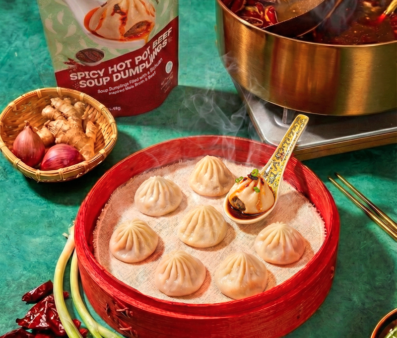 Top view of spicy hot pot beef soup dumplings showcasing pleated tops and spoonful of broth with packaging and hot pot behind